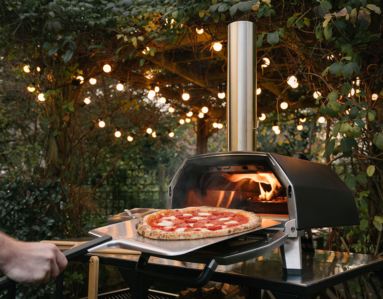 A pizza sliding into a lit outdoor pizza oven under festoon lights